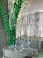 Four tall clear glass vases displayed on a shelf with green decorative sticks inside one vase and beside them.