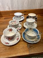 Photo showing six different teacups with matching saucers arranged in two rows on a wooden surface, each cup featuring a distinct floral design and gold accents.