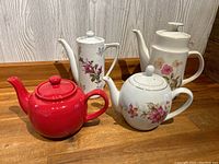 Photo of four vintage teapots, three with floral patterns and one solid red, all with lids intact on wooden surface