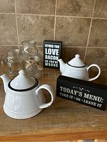 Photo showing two white ceramic teapots, two black quote signs, and two clear glass vases with twine on a wooden surface.