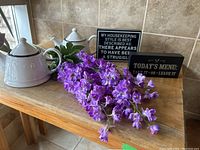 Lot contents displayed on wooden surface with tiled wall background; shows the two white ceramic teapots, two black and white quote signs, and a bunch of purple artificial delphinium flowers.