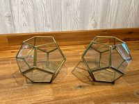 Both terrariums shown together on a wooden surface, displaying their geometric shape and brass-colored metal edges.