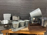 Wide view showing various metal and tin plant pots and planters including galvanized pots, decorative embossed planter, rusted rectangular planter, and metal tubs, displayed on a wooden surface against a dark tiled wall.