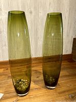 Front view of two tall green glass vases side by side, showing clear glass base and pebbles inside.