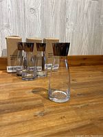 Seven mini carafe-shaped glass vases with pewter-colored bands on top, three still in original packaging boxes, arranged on a wooden surface against a light wood panel background.