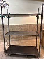 Front view of the metal wheeled utility cart showing three black board shelves, metal frame, and four wheels.