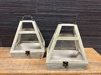 Front view of two trapezoidal wooden and glass terrariums side by side, showing metal clasp and hinged lids