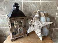 Front and side view of rustic wooden lantern with metal top, glass panels, and rattan balls inside, placed on wooden surface in front of tiled wall alongside birch wood star and glass vase