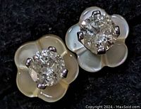 Close-up of a pair of flower-shaped earrings with mother of pearl petals and central round diamonds set in 14K white gold, displayed on a black textured background.