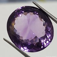 Close-up photo of oval faceted amethyst gemstone showing rich purple color and faceting detail held by tweezers.