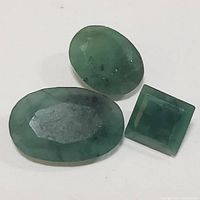 Three emerald gemstones on a white background showing oval, round, and square cuts with green coloring and visible inclusions.