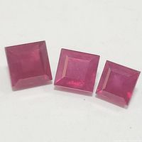 Three square-cut genuine rubies arranged on a white surface, showing red-pink hues and faceting details.