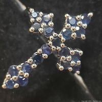Close-up of the sapphire cross design of the ring showing multiple round faceted blue sapphires set in sterling silver prongs on a black background.