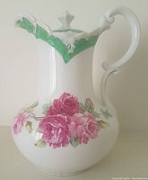 Front side view of the white porcelain antique teapot with pink and red rose floral design on the body, green and white raised accents on spout and rim, ornate curved handle, and lid finial.