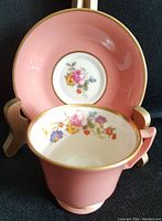 Close-up photo of demitasse cup showing pink exterior and floral interior with gold trim, displayed with matching saucer on stand.