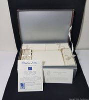 Opened original silver box showing four tied bundles of small envelopes around a stack of stationary paper in the center, with two printed informative inserts included.