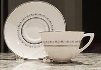 Front view of one teacup with matching saucer set behind. White porcelain with silver floral pattern band and silver rim.