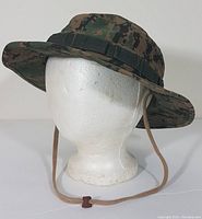 Front-left angled view of the camouflage safari hat showing wide brim, loops around the crown, and chin strap with toggle.