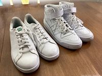 Two pairs of white Adidas sneakers placed side by side on a wooden floor, showing both low-top and high-top styles.