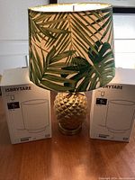 Front photo showing a golden pineapple base lamp with a tropical leaf print fabric lampshade placed between two original boxed IKEA ISBRYTARE table lamps.