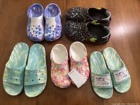 Top view showing five pairs of Crocs: blue/white tie dye slides, black splatter Velcro clogs, multi-color tie dye new clogs with tags, blue/purple/white clogs, and Jon Cho slides with cartoons.