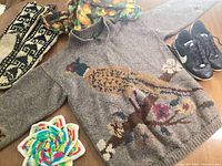 Wool sweater with mallard bird design, knitted moccasins, Nike Cortez shoes, orange patterned skirt, and a multicolor knitted trivet laid out on a table.