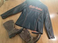 Full view of Harley-Davidson black leather jacket, women's size XS, with orange logos and piping, paired with brown leather Harley-Davidson boots, size 7, showing general condition of items.