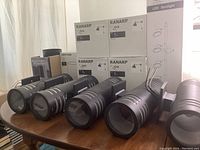 Five black cylindrical exterior sconce lights with frosted glass strips on sides, displayed on wood table in front of RANARP and LED spotlight light boxes.