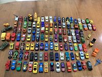 Top-down view of about 73 assorted diecast toy cars arranged neatly in rows on a wooden surface showing variety of colors, styles, and sizes.