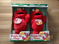 Two Tickle Me Elmo dolls in original green and white boxes, front view showing the dolls and Try Me button.