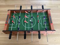 Top-down image of mini foosball table showing green playing field with player figures in red and blue, black rubber handles, and mini soccer ball in play area.