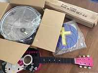 Photo showing packed snare drum partially in box, hi-hat stand box, packaged tambourine, and Hello Kitty acoustic guitar lying flat