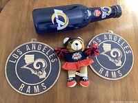 Lot items arranged on wooden surface: LA Rams blue bottle bank, Super Bowl XXXIV cheerleader teddy bear plush, and two round wood Rams wall signs.