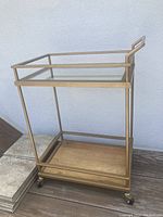Full view of the gold bar cart showing metal frame, glass top shelf, and wood bottom shelf.