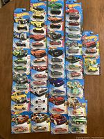 Photo displaying 38 Hot Wheels cars in original packaging arranged in rows on a wooden surface, showcasing various models, colors, and some multi-packs.