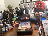 Photo showing multiple Star Wars action figures including Stormtroopers, Darth Vader, Luke, Leia, Yoda, Anakin; black Star Wars bank figure; Kylo Ren bobblehead in packaging; Star Wars pinball handheld game; two puzzles in boxes; small Star Wars accessories like candy dispensers, watch, keychain, and Band-aid container; Star Wars themed backpack in background.