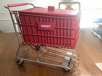 Side angled view of a red miniature Target shopping cart on a wooden surface showing the metallic frame, red basket with Target logo, and four wheels.