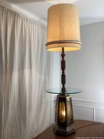 Full view of the vintage floor lamp showing the tall wooden turned central column, large beige cylindrical fabric shade with wear, clear round glass tabletop attached at mid-height, and octagonal metal base with amber hammered panels.