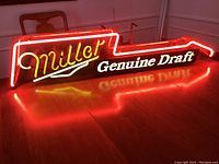 Front view of illuminated vintage Miller Genuine Draft neon sign shaped like a guitar, showing red and yellow neon tubing with white illuminated letters and reflection on wood floor.