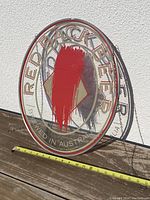 Front view of vintage circular Red Back Beer glass hanging sign leaning against white wall with measuring tape for scale, approx 13 inches diameter.