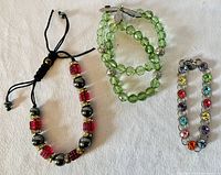 Three bracelets laid out on white cloth: black cord bracelet with red crystals and metallic beads, green glass bead bracelet with a clasp, and multicolored rhinestone bracelet on metal chain.