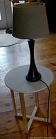 Side view of table with white round top, wood legs in natural finish, and dark tone table lamp with beige shade on it.