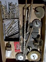 Overview of over 40 drill bits in various sizes arranged in metal container and scattered in box, alongside several metallic grinding and sanding attachments including wire brushes and abrasive discs, plus a long large hole saw bit with white base.
