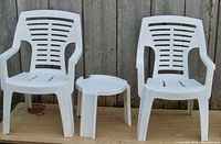 Front view of the outdoor patio set showing two white resin armchairs and a small matching white resin end table on a wooden surface against a wooden fence.
