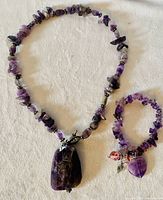 Photo of full necklace and bracelet set laid flat on light background, showing amethyst stone beads and large rectangular pendant on necklace, and purple amethyst chip beads with charm pendants on bracelet.
