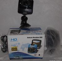 Photo of black square dash camera mounted on suction cup mount, placed on top of original product box labeled 'Vehicle Blackbox DVR' with HD and Full HD 1080 logos. Cables and plastic-wrapped accessories visible to the side.