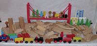 Wooden train set pieces arranged featuring multiple curved and straight tracks, a red suspension bridge, colorful wooden trains with wheels, trees, people figures, various signs, and background scenery pieces.