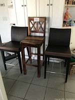 Three bar chairs: two black faux leather chairs with stainless steel footrests, and one solid wood bar chair with cross-back, against a white cabinet background.