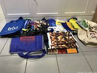 Lot of assorted reusable bags laid out on floor, showing various sizes, colors, and printed logos including blue, beige, and multicolored bags.