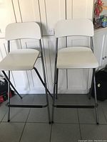 Two white bar chairs with black metal legs standing side by side against a white wainscoted wall.